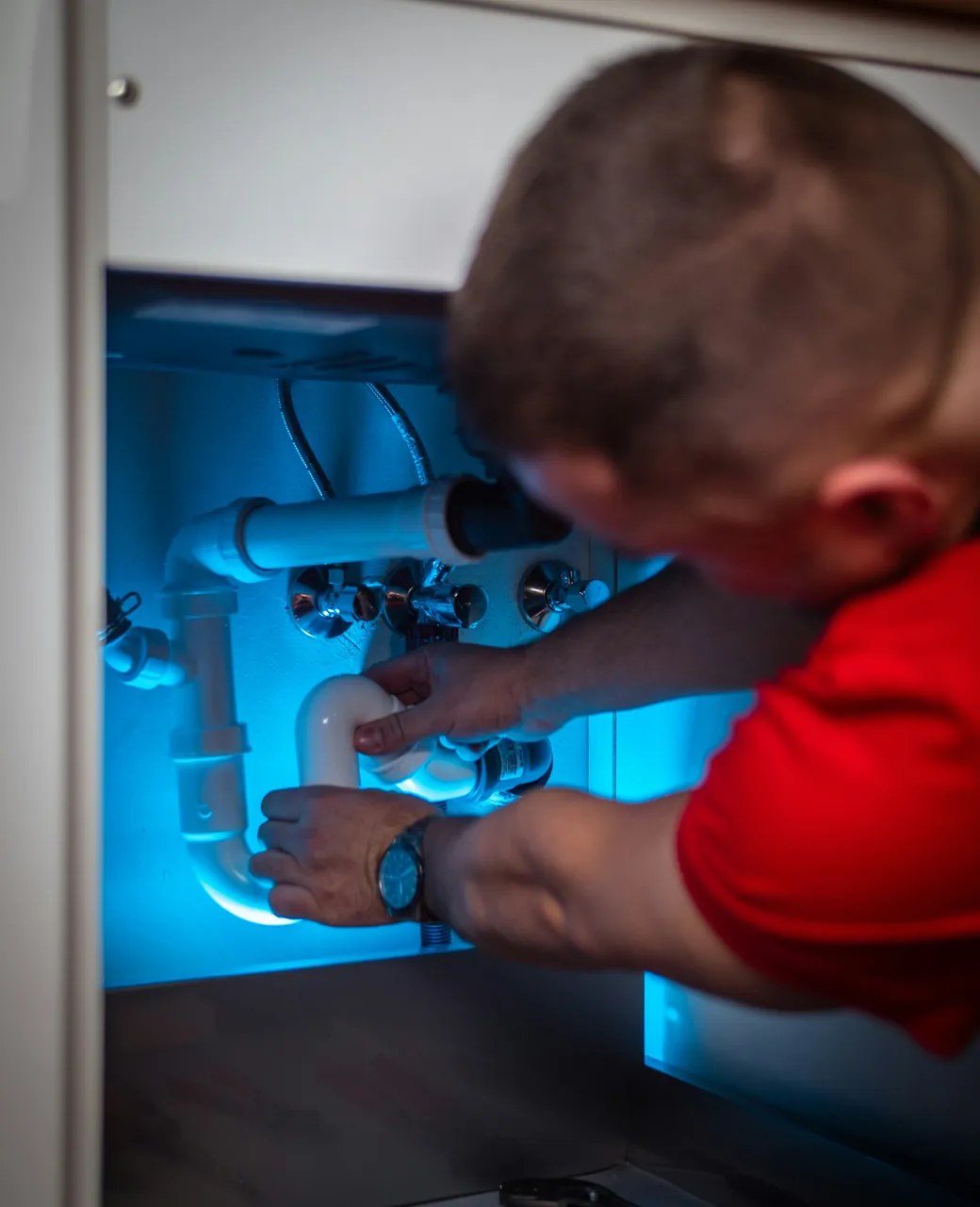 Plombier en polo rouge réparant le siphon d’un évier dans un meuble éclairé en bleu